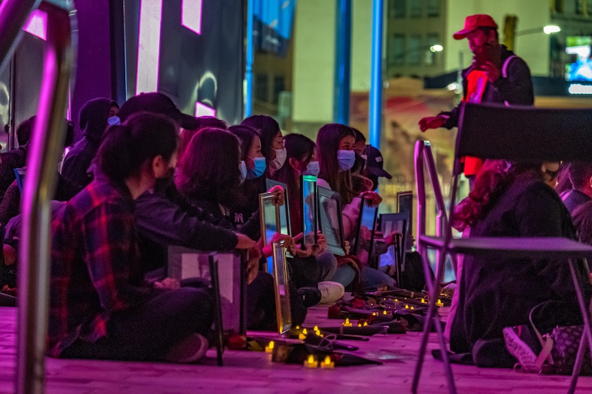 Women in masks hold framed photographs and kneel at a vigil with candles in a purple dark night. 