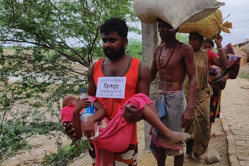 A volunteer carries a sick child as villagers walk in a line carrying goods on their heads.