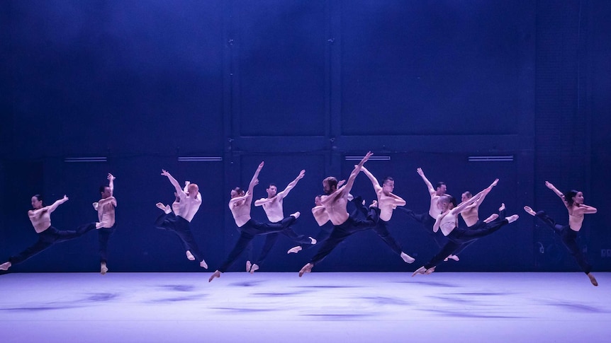 Thirteen dancers, both male and female, leap across a white stage in front of a dark blue backdrop.