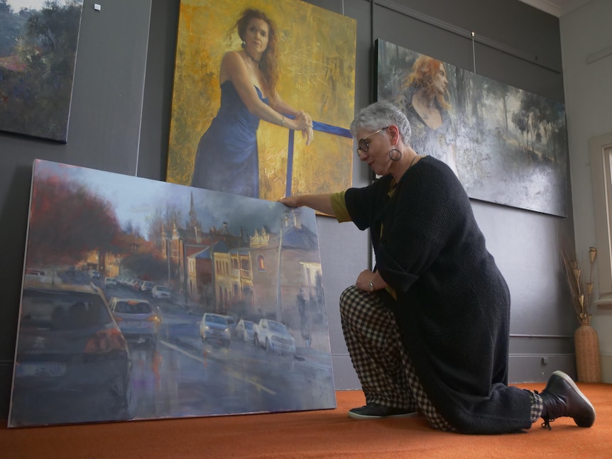 A woman kneeling on the floor while she looks at a large streetscape painting.
