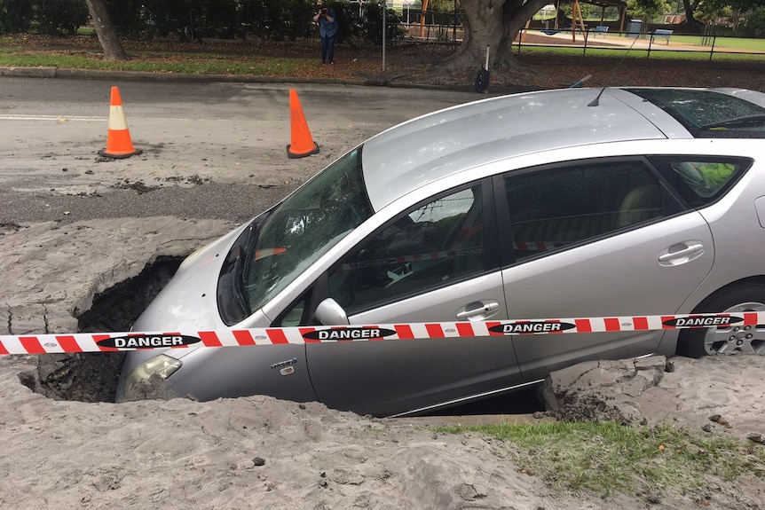 A car rests in a large hole in a roadway.