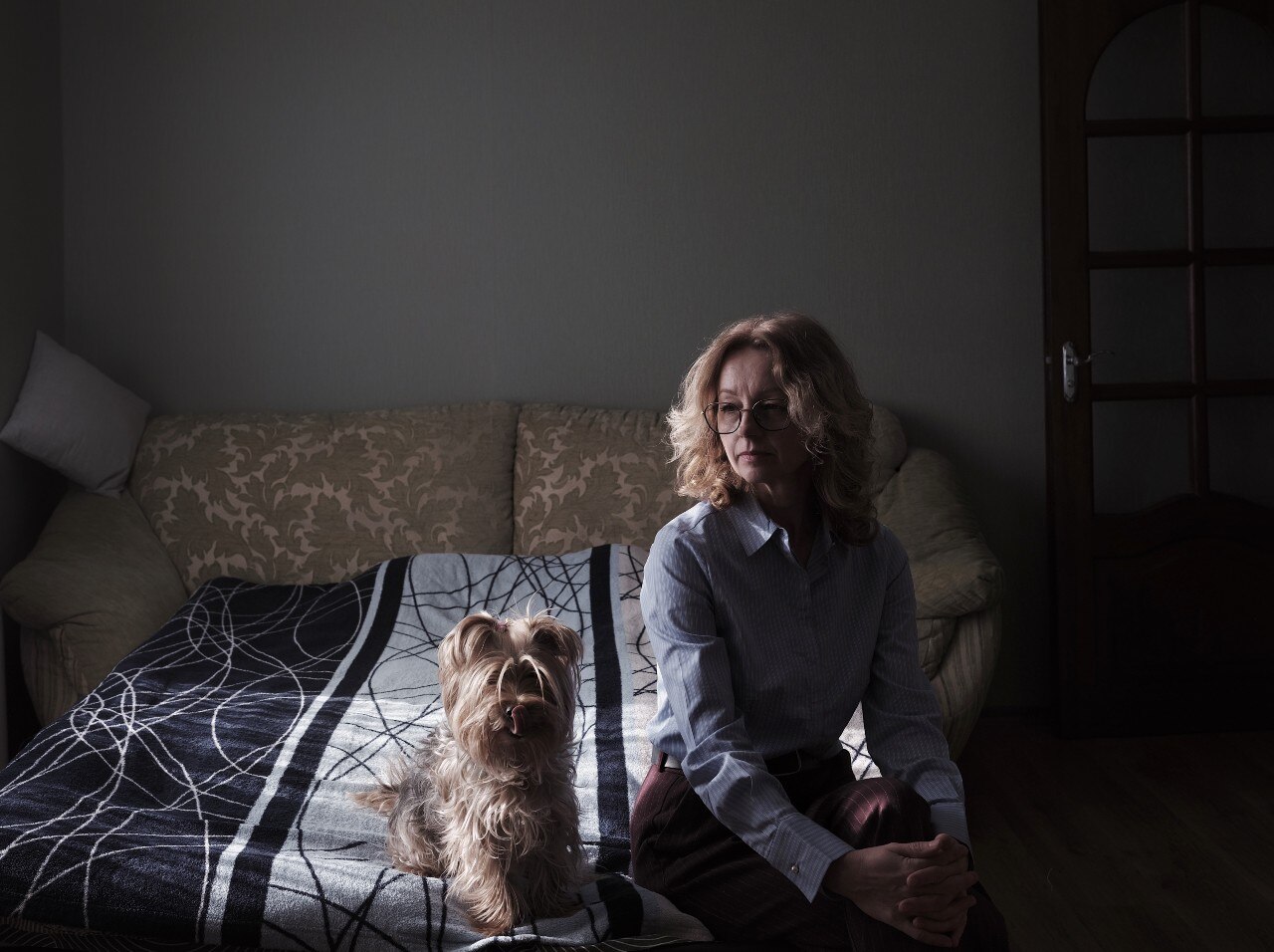 A woman sitting on a bed, looking on. A dog is also visible.