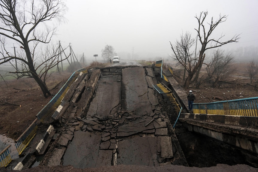 A man walks along a broken bridge buckled by bombings, with trees bent over in the fog
