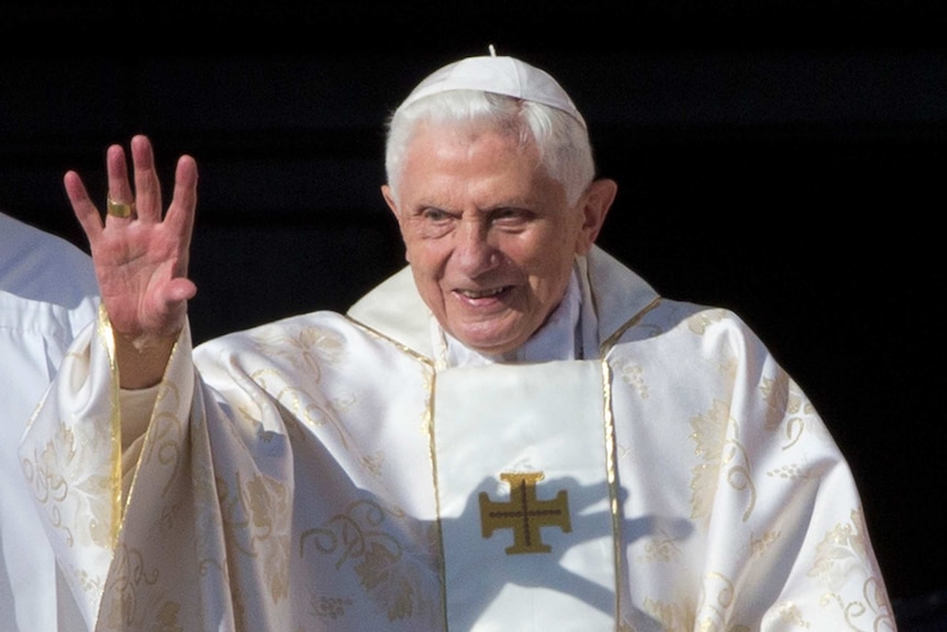 Pope Benedict waves to a crowd while wearing a white cap and white robes.