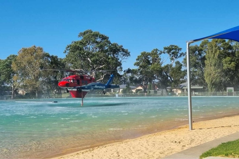 A helicopter hovers over a man-made lake suctioning water next to a beach and gazebo