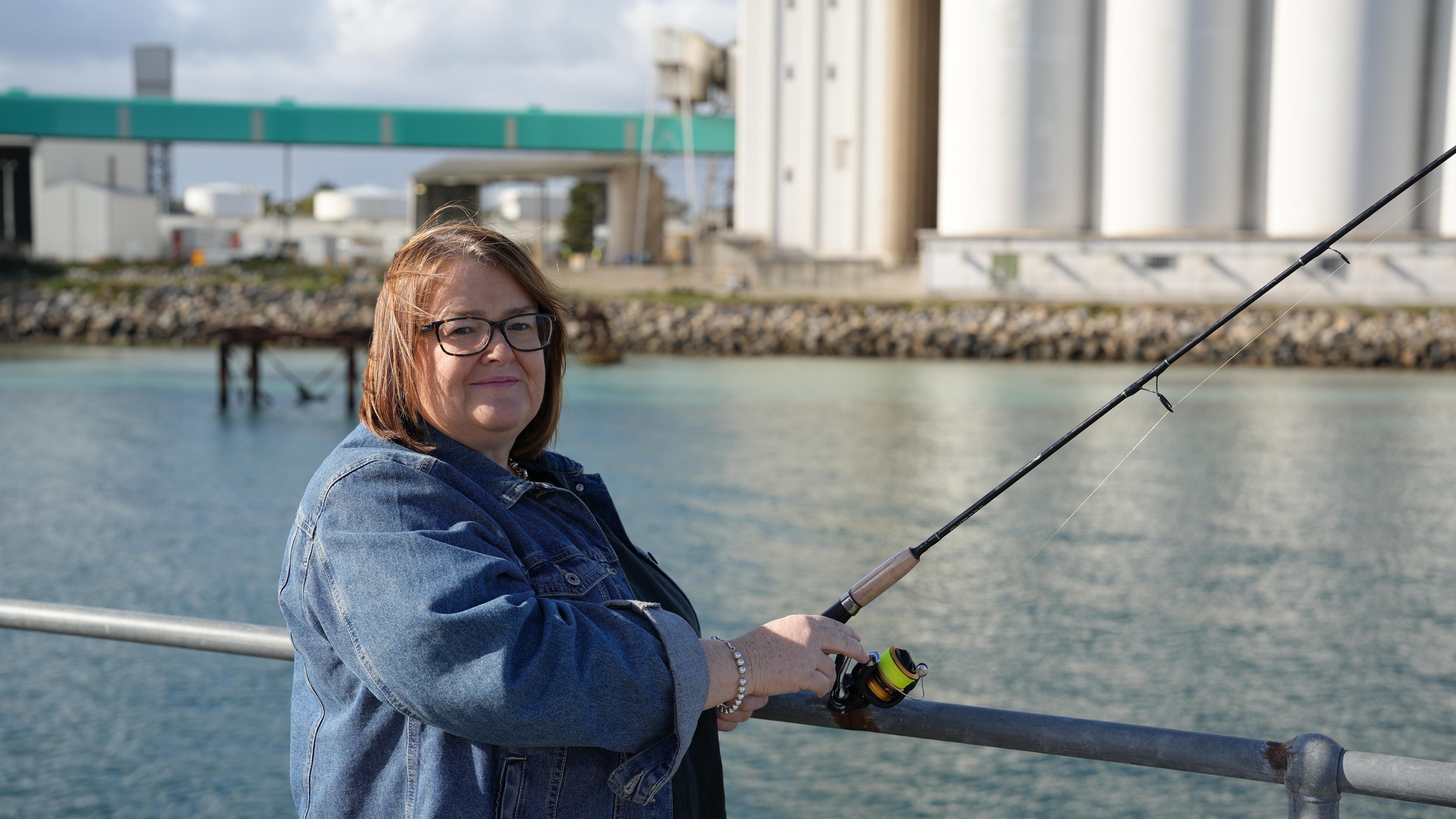 A woman stands on a jetty holding a fishing rod, ready to cast her line into the water.
