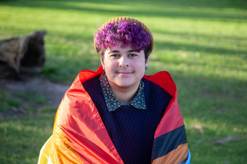 A man with purple hair wraps himself in a pride flag