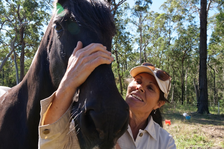 A woman stands with her arm around her horse, with their heads together.