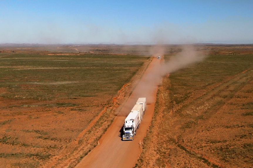 Large red dirt landscape with dirt track road. Road train drives on track kicking up dust clouds. 