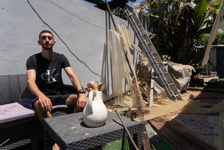 A young man sits outside, with damage to surrounding buildings visible