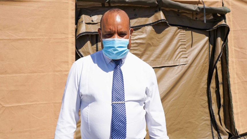 A Papua New Guinean man in a white shirt, tie and medical face mask stands in front of a brown tent