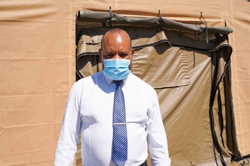 A Papua New Guinean man in a white shirt, tie and medical face mask stands in front of a brown tent