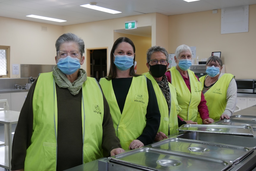 five women in yellow hi-vis vests stand together along a commercial kitchen bench lined with a row of food warmers