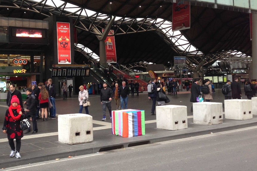 A series of large concrete blocks outside a busy railway station, one covered in stripy colourful material.