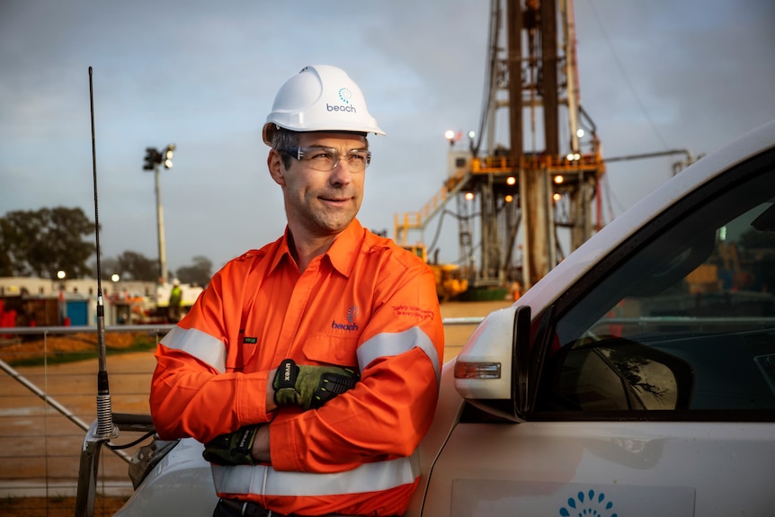 Man wearing hard hat and protective glasses, orange high-viz shirt, stands in front of gas drilling rig, arms crossed, smiles.