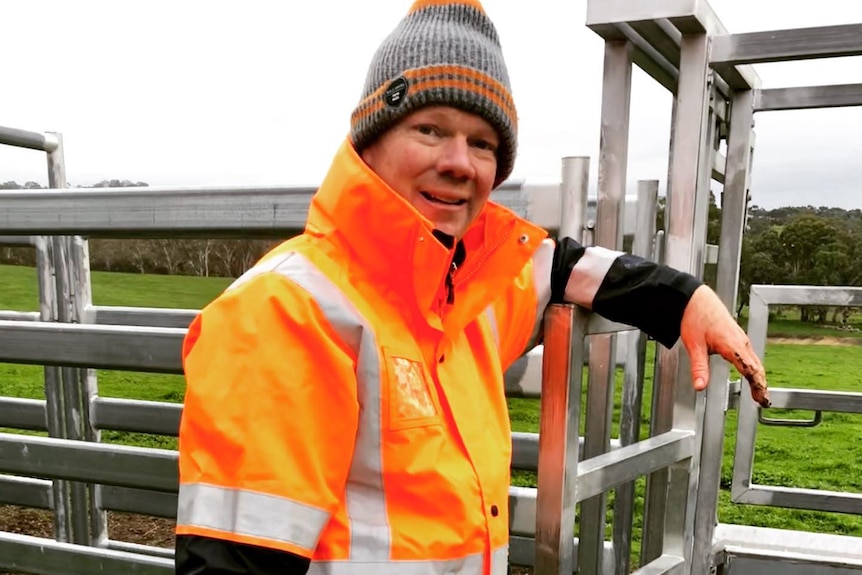 A smiling farmer with a high visibility orange jacket and a gray beanie on a farm.