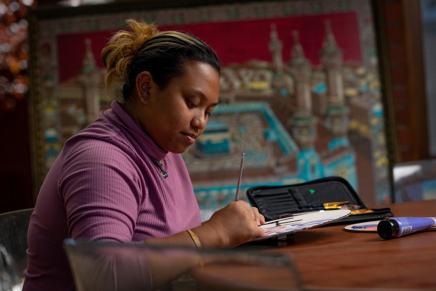 A woman sits at a table painting with art supplies on the table and a large artwork in the background