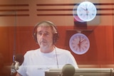 A middle-aged man with a grey goatee beard stands in the radio studio, amid several clocks reflected in the glass
