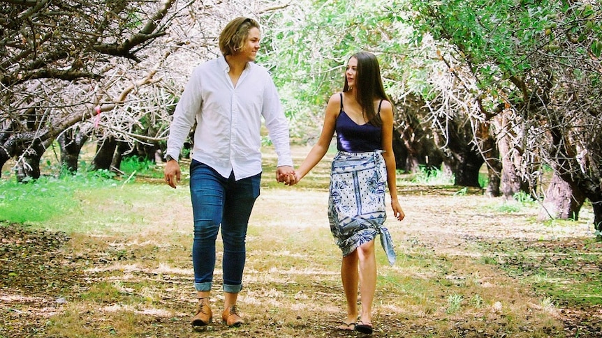 A couple walks through a grove of trees, holding hands