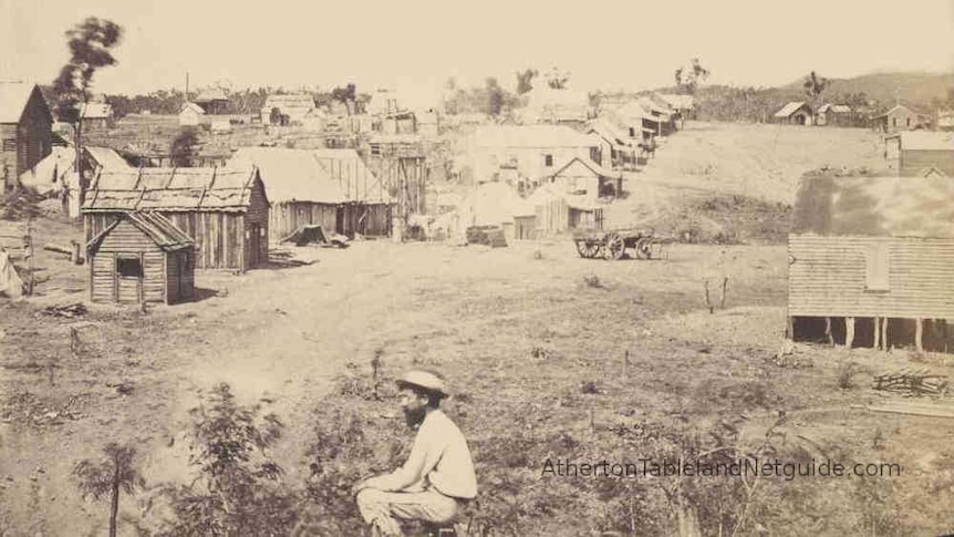 A black and white photo of a shanty town along a dusty road with a man sitting in the foreground.