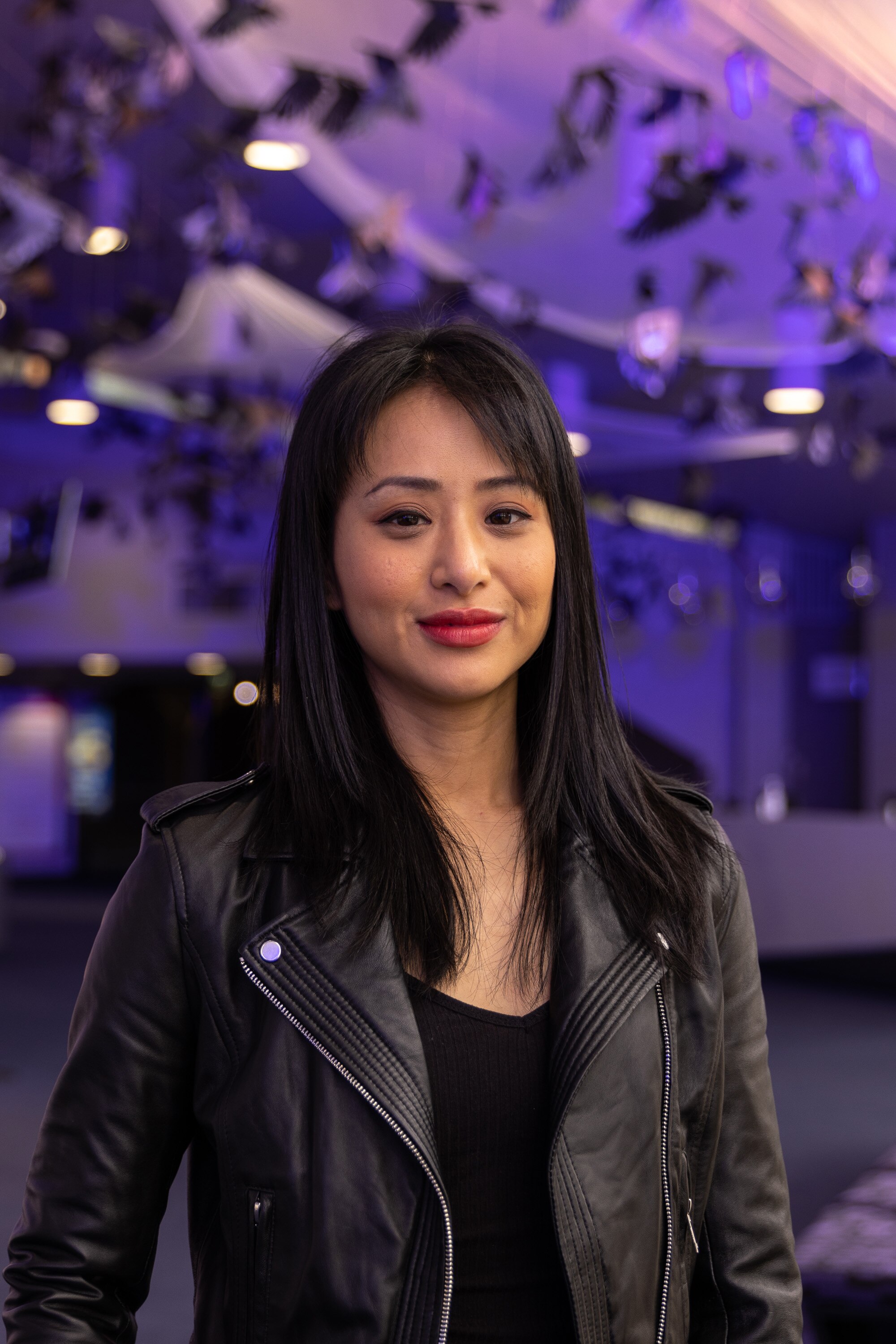 An Asian Australian woman with a fringe, wearing a leather jacket, stands in a theatre foyer. She smiles slightly at the camera.