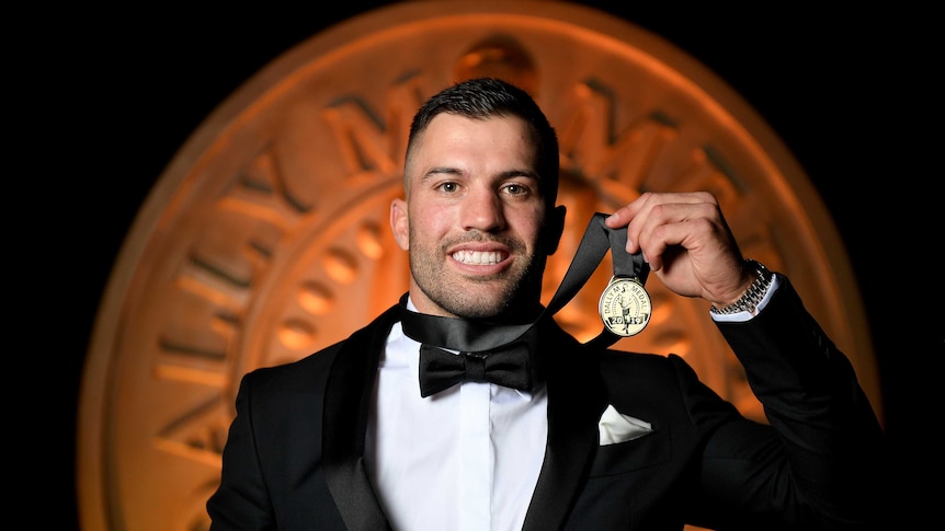 James Tedesco holds a medal with a black ribbon towards the camera in front of a Dally M logo.