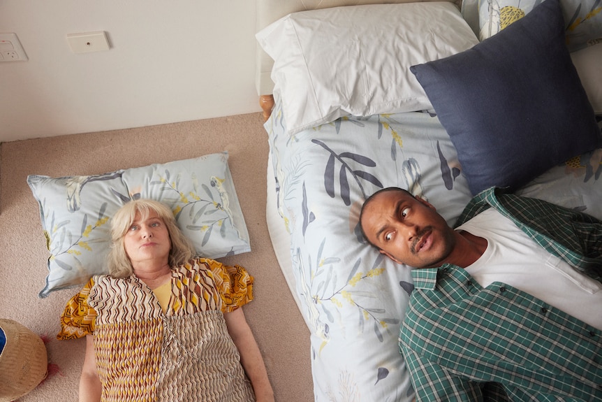 Matt Okine in Mother and Son, lying on a bed, while his TV mum Denise Scott lies on the floor next to him, staring into space.