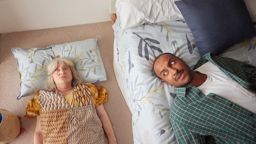 Matt Okine in Mother and Son, lying on a bed, while his TV mum Denise Scott lies on the floor next to him, staring into space.