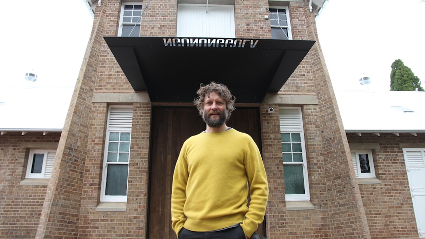 Ben Quilty wears a yellow jumper and stands out the front of the gallery's entrance.