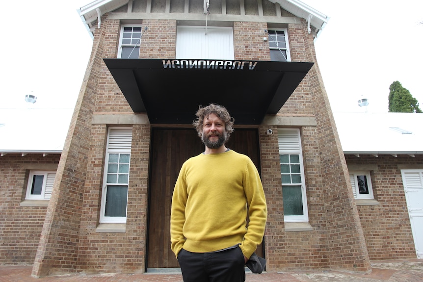 Ben Quilty wears a yellow jumper and stands out the front of the gallery's entrance.
