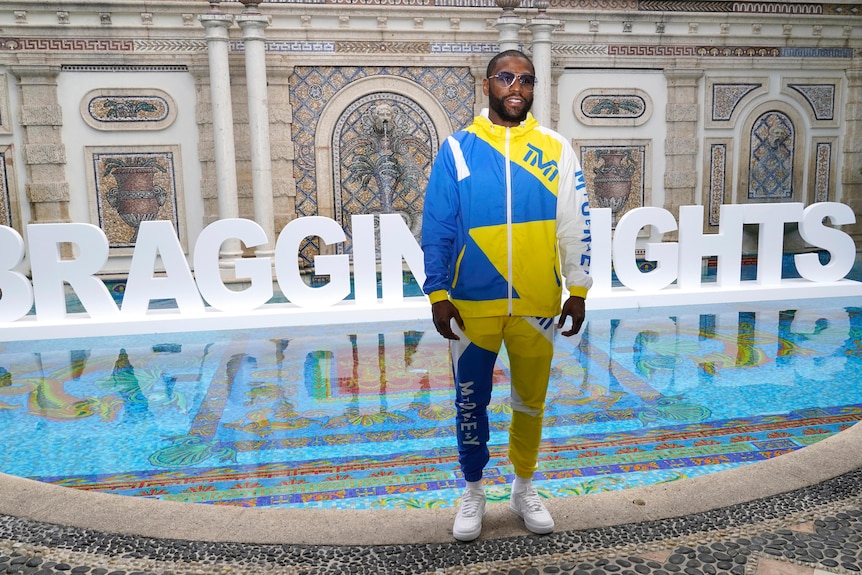 Superstar boxer Floyd Mayweather poses in a blue and yellow tracksuit, with a sign behind him that says "Bragging Rights". 
