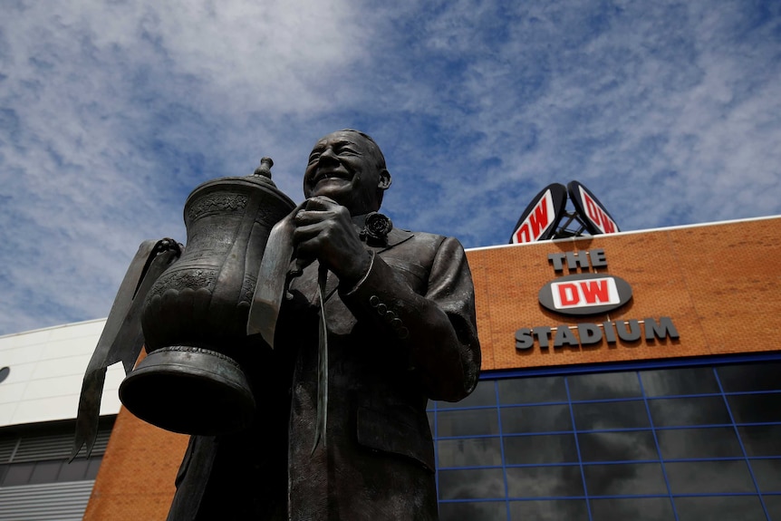 A statue of a man holding the FA Cup is pictured from the base looking up at a blue sky with the outside of a stadium in shot