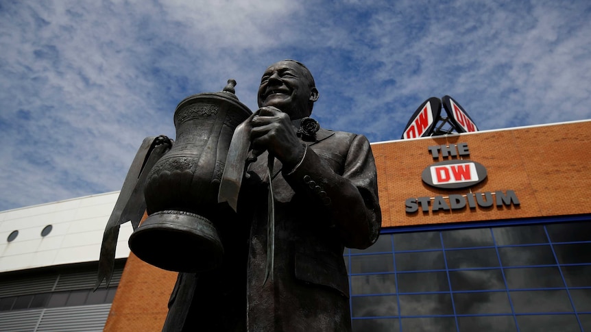 A statue of a man holding the FA Cup is pictured from the base looking up at a blue sky with the outside of a stadium in shot