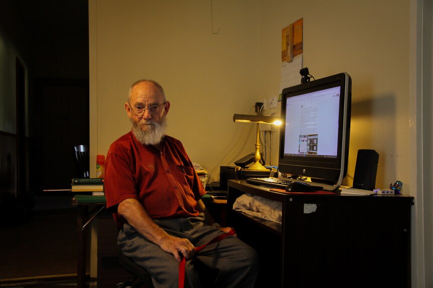 An elderly man with a grey beard sitting a desk in front of a computer while looking at the camera.