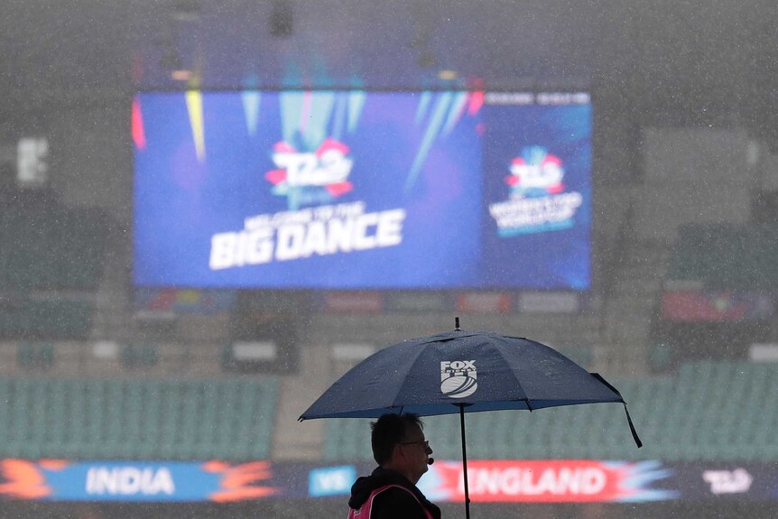 A man with a Fox Sports umbrella stands in the rain at a stadium while the big screen shows "The Big Dance" in the background.