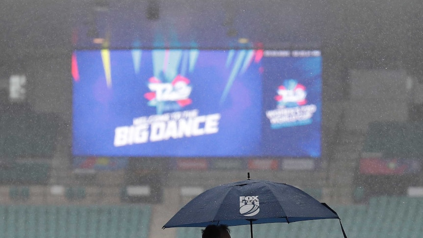 A man with a Fox Sports umbrella stands in the rain at a stadium while the big screen shows "The Big Dance" in the background.