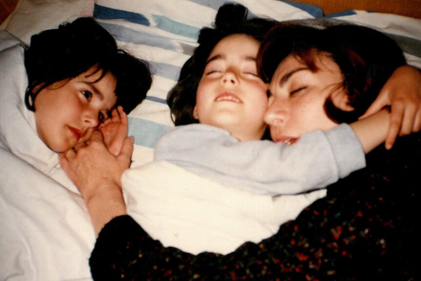 An old family photo of two girls lying in bed cuddling their mother.