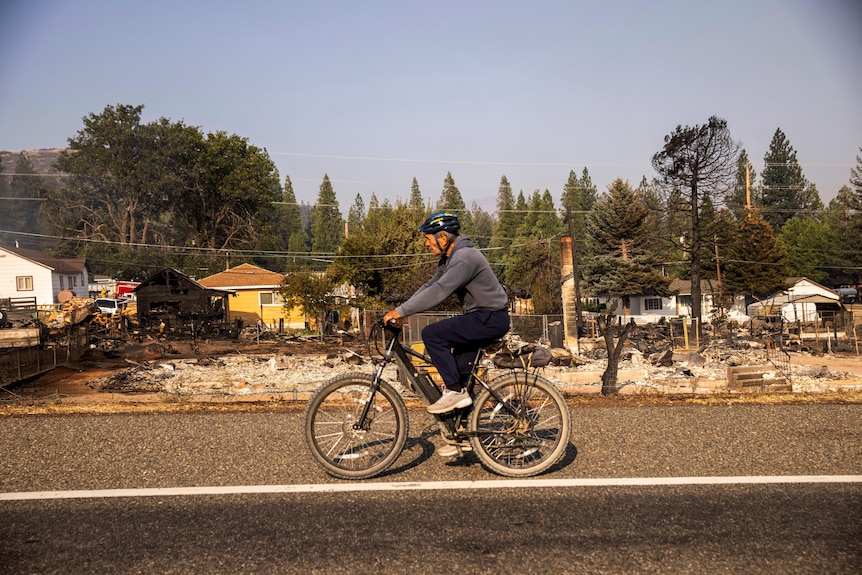 Un uomo va in bicicletta sulla strada.  Sullo sfondo, le case vengono distrutte da un incendio. 