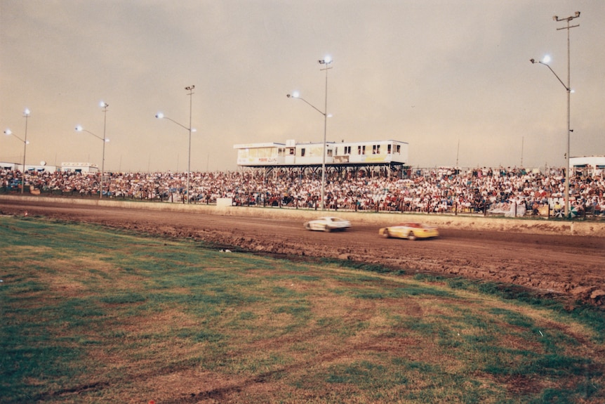 Crowds pack the hills are two cars zoom along the dirt oval track of a speedway.