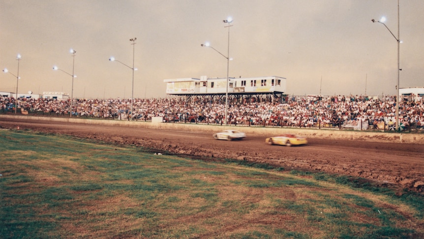 Crowds pack the hills are two cars zoom along the dirt oval track of a speedway.