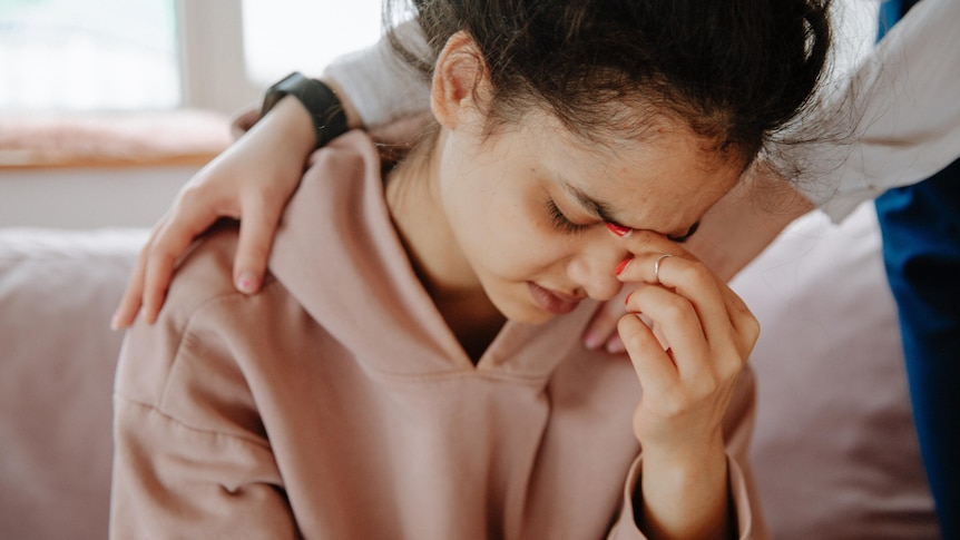 A young woman holding her face in her hands, as someone comforts her.