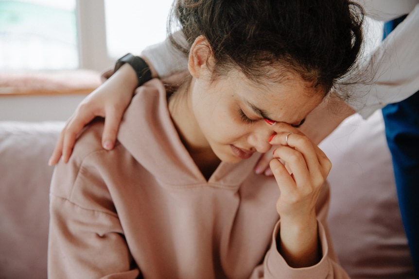 A young woman holding her face in her hands, as someone comforts her.