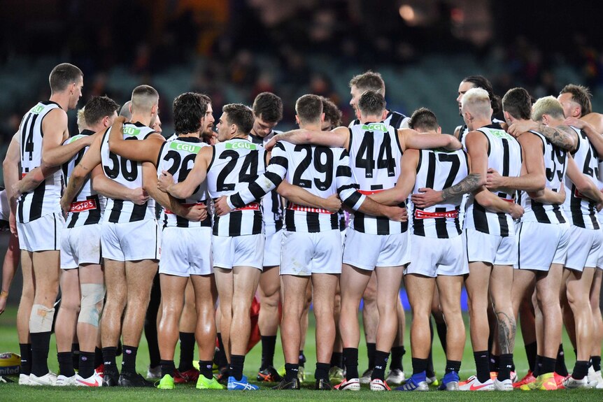 A huddle of AFL players wearing black and white jerseys at a night-time match in half-full stadium.