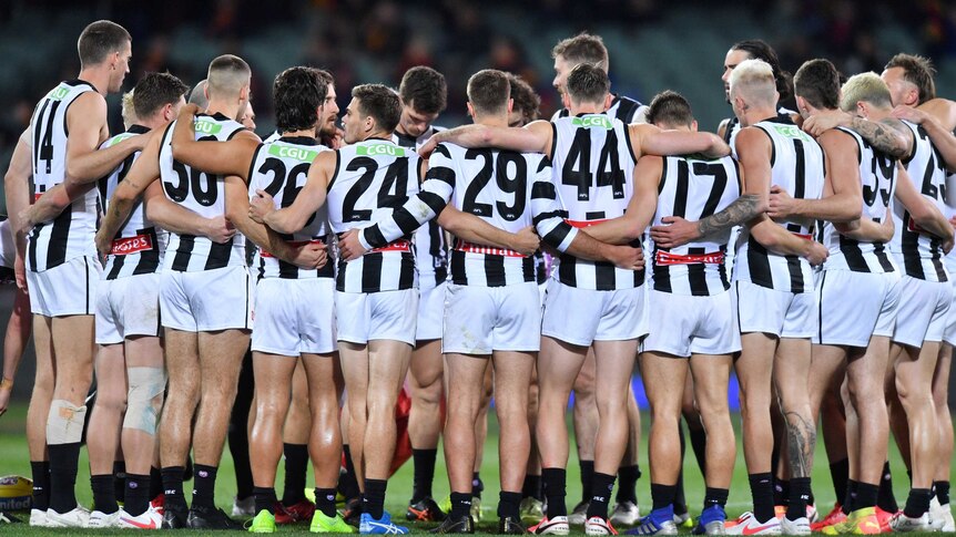 A huddle of AFL players wearing black and white jerseys at a night-time match in half-full stadium.