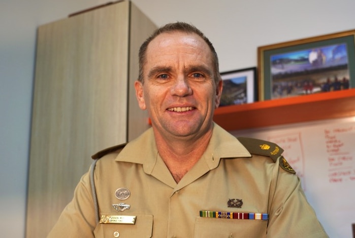 A late middle-aged man in military dress sits at a desk, holding a pen and smiling.