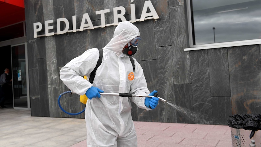 A man in a white biohazard suit and black mask sprays disinfectant outside a Spanish hospital.