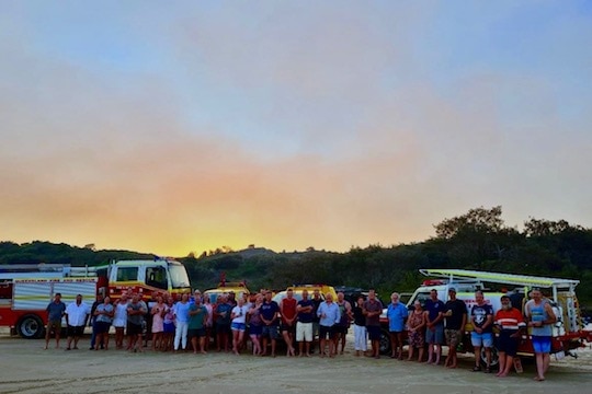 A group on a beach with fire trucks and smoke.