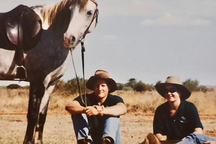 Man and woman wearing hats sitting on the ground in front of a grey horse. The man is holding the reins.