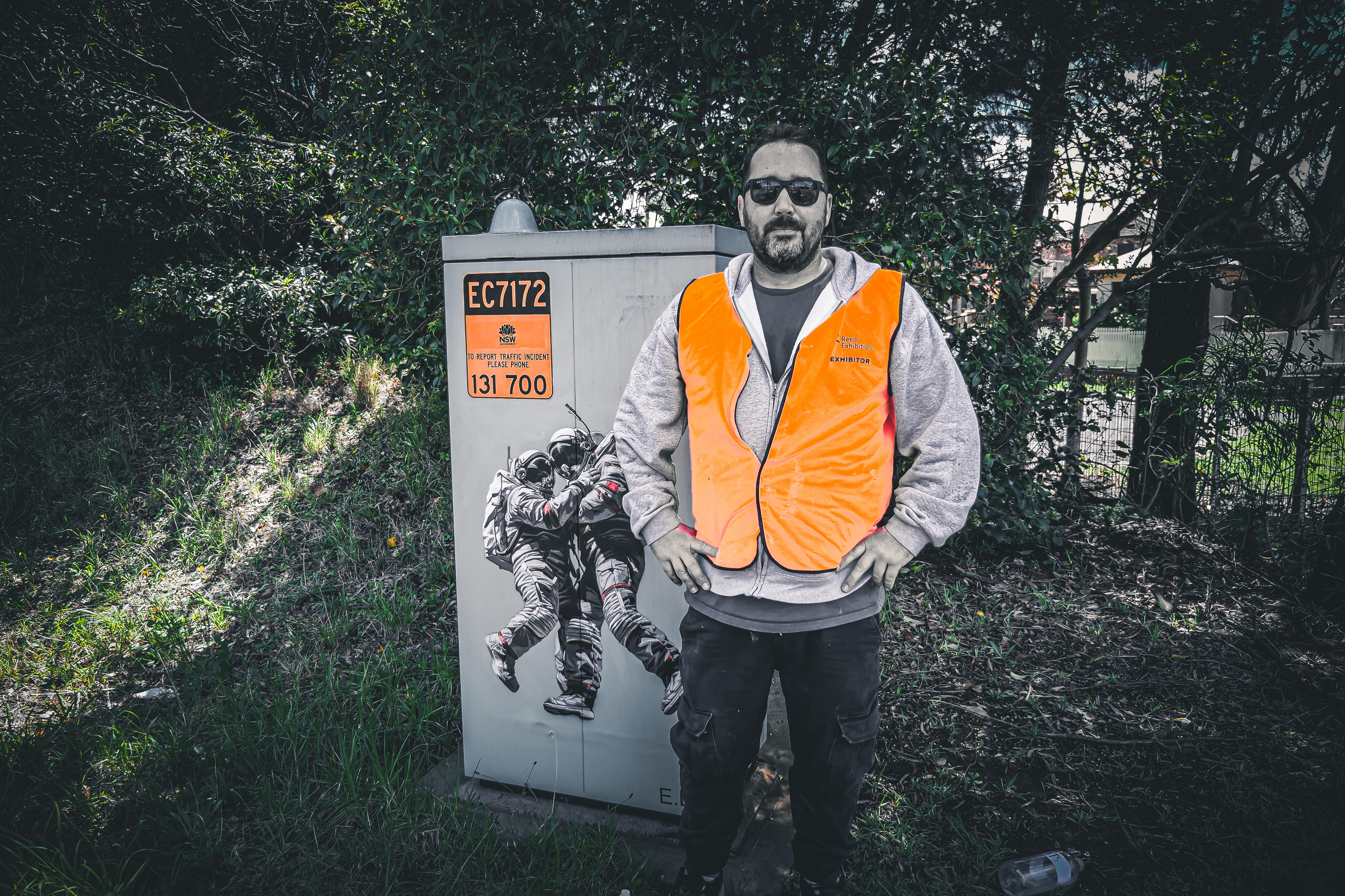 A man wearing a fluro jacket stands beside a painted traffic control box