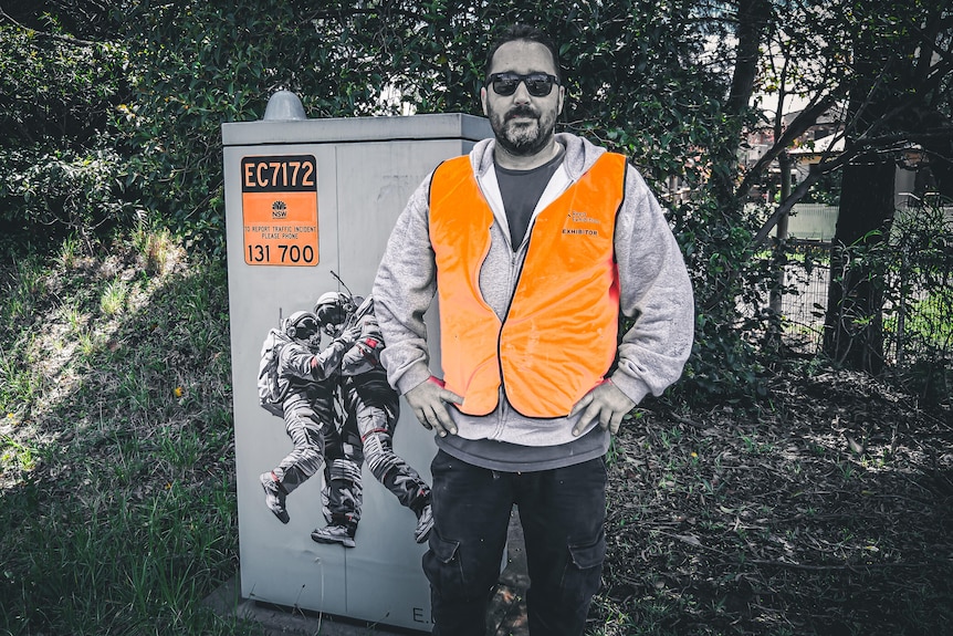 A man wearing a fluro jacket stands beside a painted traffic control box